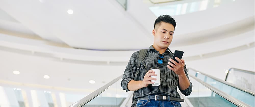 man holding coffee cup looking down at phone while riding down escalator