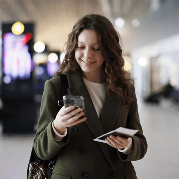 молодая женщина смотрит в экран телефона с паспортом и билетом в руке