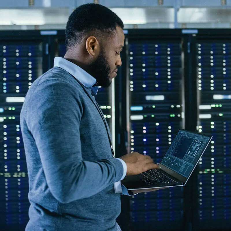 hombre mirando una computadora mientras está de pie en una sala de servidores vistiendo un conjunto de suéter y camisa