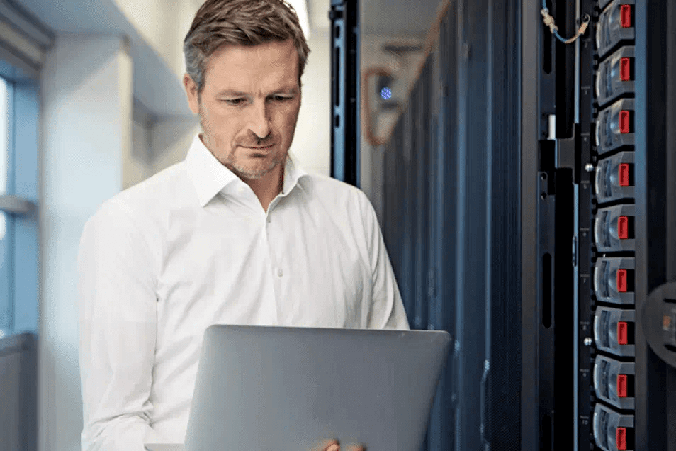 man with white shirt looking down at laptop in server room