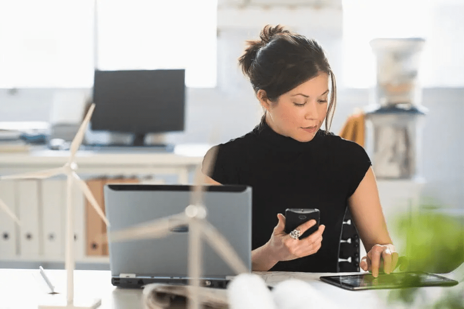 Frau mit Telefon in der Hand sitzt vor Computer und tippt auf Tablet