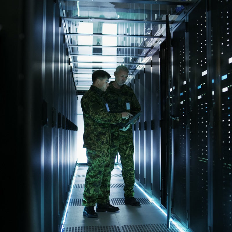 In Data Center Two Military Men Work with Open Server Rack Cabinet. One Holds Military Edition Laptop.