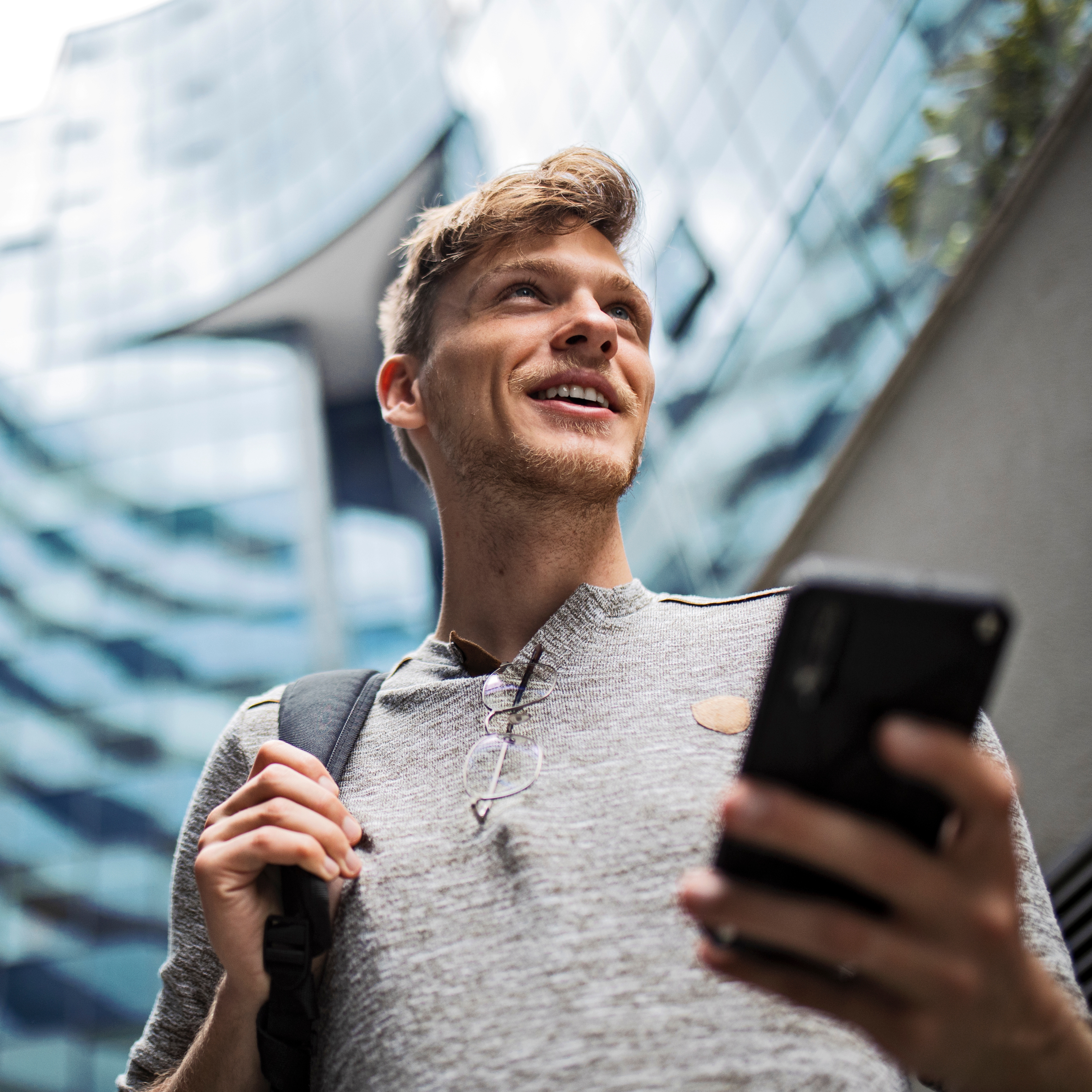 Personne tenant un smartphone et portant un sac à dos, debout en plein air près de bâtiments modernes en verre