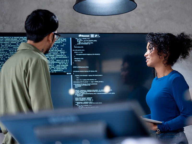 Zwei Personen stehen vor einem großen Bildschirm, auf dem Code angezeigt wird, und diskutieren über Softwareentwicklung