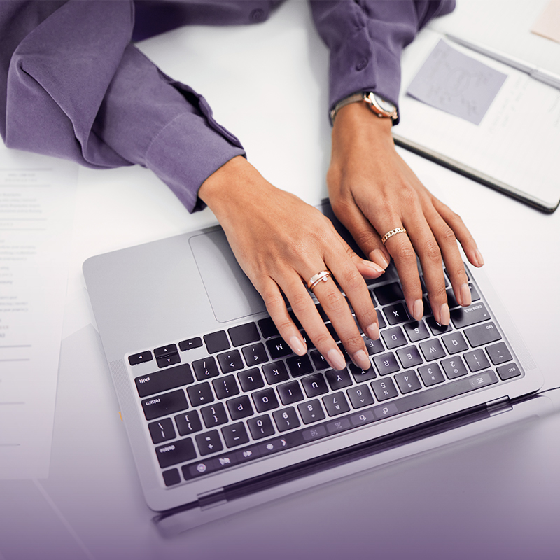 Hands typing on a laptop keyboard at a desk with a notebook nearby
