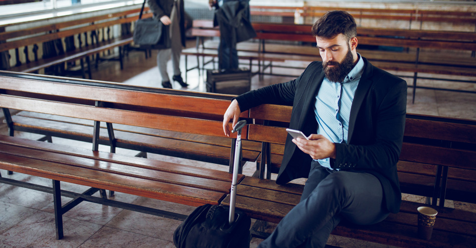 Person sitting on a bench in a transit station, looking at a phone with a suitcase beside them