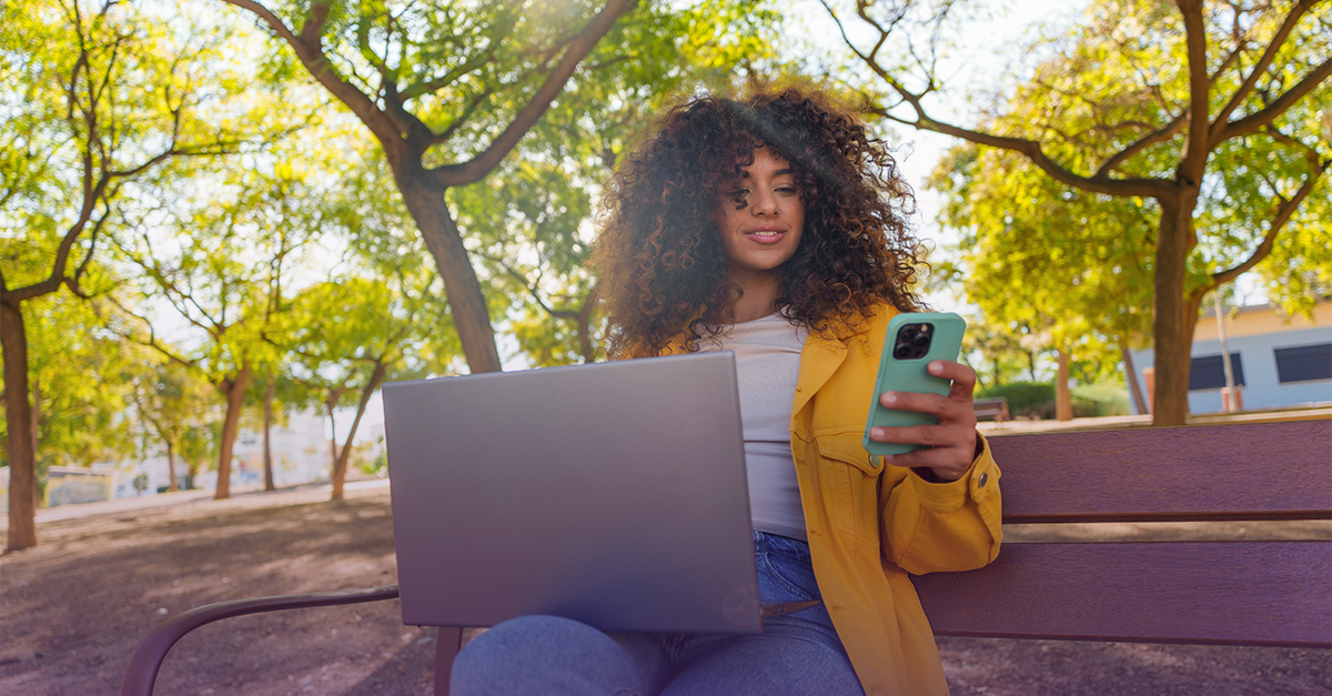 Person sitting on a park bench, using a laptop while holding a smartphone outdoors among trees