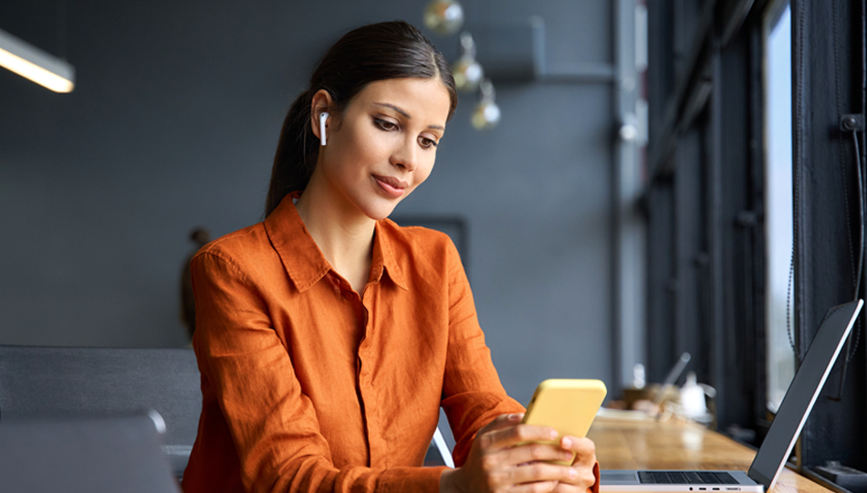 Person using a smartphone while sitting at a desk with a laptop in a modern workspace